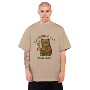 Peut inclure: Un t-shirt beige avec une illustration d'ours en colère et le texte "THE WORLD CAN WAIT". L'ours tient une tasse de café avec un crâne. Le t-shirt a un col rond et des manches courtes. L'ours est assis à une petite table.
