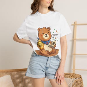 Peut inclure: T-shirt blanc avec un ours de dessin animé tenant un ordinateur portable et un sac d'argent. Le texte "SUCCESS!" est au-dessus. Le sac d'argent porte l'inscription "CERTIFIED: Works On My Machine". L'ours porte un bandana bleu.