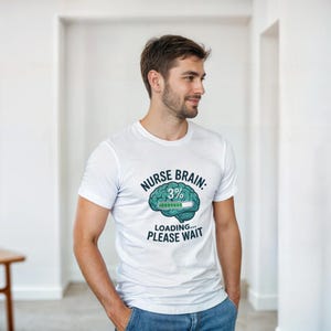 Peut inclure: T-shirt blanc avec un graphisme humoristique. Le motif représente un cerveau turquoise avec une barre de chargement à 3% et le texte "NURSE BRAIN: 3% LOADING... PLEASE WAIT". Le t-shirt est porté par une personne en jean bleu.
