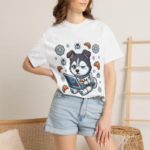 Può includere: T-shirt bianca con un design di cane husky pixelato che utilizza un laptop. Il cane indossa una tuta spaziale ed è circondato da fiocchi di neve, croissant e insetti. Il testo sullo schermo del laptop è visibile.
