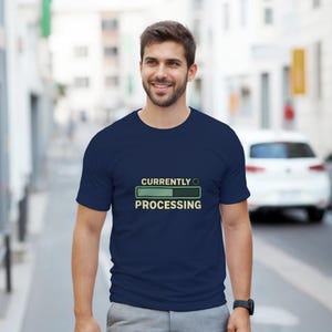 Puede incluir: Una camiseta azul marino con el texto "CURRENTLY PROCESSING" encima de una barra de progreso verde. La camiseta la lleva una persona sonriendo al aire libre.