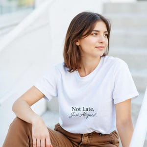 Peut inclure: Un t-shirt blanc avec le texte "Not Late, Just Aligned" en écriture cursive. Le t-shirt est porté par une personne assise sur des marches, portant un pantalon marron. L'esthétique générale est décontractée et détendue.