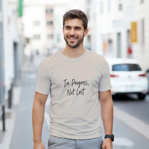 Peut inclure: Un t-shirt gris clair avec le texte manuscrit "In Progress, Not Lost" en noir. Le t-shirt est porté par une personne souriante en extérieur. La personne porte une montre noire.