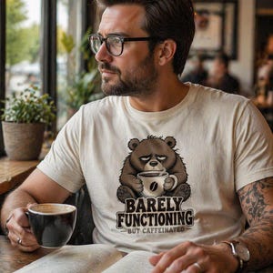 Peut inclure: Un t-shirt à manches courtes de couleur crème avec un motif représentant un ours brun fatigué tenant une tasse de café. Sous l'ours, le texte indique BARELY FUNCTIONING en grandes lettres et BUT CAFFEINATED en plus petit, avec un grain de café.