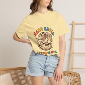 May include: Yellow t-shirt with a cartoon armadillo illustration. The armadillo is brown and beige, curled up with the text "HARD SHELL SOFT CRISIS INSIDE" in a rainbow font. The shirt is a light yellow color.