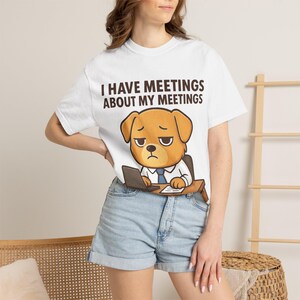 Peut inclure: T-shirt blanc avec l'inscription "I HAVE MEETINGS ABOUT MY MEETINGS" et un dessin de chien de dessin animé portant une cravate et travaillant sur un ordinateur portable. Le chien a une expression triste. Le t-shirt est porté avec un short en jean.