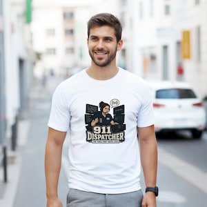 Peut inclure: Un t-shirt blanc à manches courtes avec une impression graphique représentant un répartiteur 911 portant un casque, assis à un bureau avec plusieurs écrans. Le texte indique 911 DISPATCHER THE VOICE THAT SAVES LIVES avec une icône de signal.