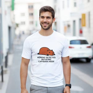 Peut inclure: T-shirt blanc avec un dessin de capybara marron et le texte "MONDAY DETECTED. INITIATING CAPYBARA MODE." Le capybara est représenté allongé. Le t-shirt est porté par une personne.