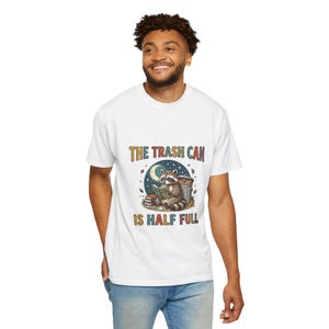 Camiseta "El cubo de basura está medio lleno" | Divertida camiseta con un mapache filósofo leyendo un libro y un mensaje de humor optimista.