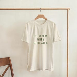 Può includere: T-shirt color crema con la scritta "I'll Return When Regulated" in verde scuro. La maglietta a maniche corte è appesa a una gruccia di legno sospesa a un attaccapanni. La maglietta è realizzata in un materiale morbido e leggero.