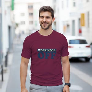 Peut inclure: Un t-shirt bordeaux avec le texte "WORK MODE: OFF" en bleu, avec une police vieillie. Le t-shirt à manches courtes est porté par une personne souriante, avec un arrière-plan flou d'une rue et d'une voiture blanche.