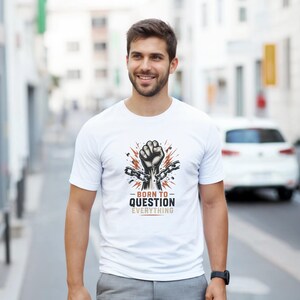 Peut inclure: Un t-shirt blanc à manches courtes orné d'un motif représentant un poing levé brisant des chaînes métalliques. Sous le poing, le texte BORN TO QUESTION EVERYTHING est inscrit en lettres noires et beiges avec un motif d'éclat stylisé.