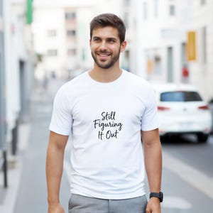 Peut inclure: Un t-shirt blanc avec le texte "Still Figuring It Out" en noir. La personne porte le t-shirt dans un cadre urbain.