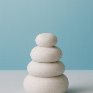 May include: A stack of four smooth, light-colored stones arranged in a tower. The stones are oval-shaped and vary slightly in size, with the largest at the base and the smallest at the top. The background is a solid light blue.