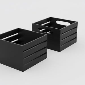 Puede incluir: Dos cajas de almacenamiento de madera negra. Una caja tiene dos asas cortadas en los lados. Ambas cajas tienen listones horizontales en los lados. Las cajas son cuadradas y se asientan sobre una superficie blanca.