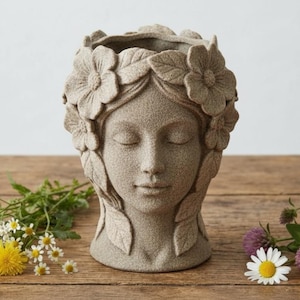 Könnte beinhalten: Ein beige-farbener, geformter Pflanztopf in Form eines Frauengesichts, verziert mit floralen und Blattdetails. Die geschlossenen Augen und der ruhige Ausdruck erzeugen eine ruhige Ästhetik. Der Pflanztopf wird auf einer Holzoberfläche mit bunten Wildblumen präsentiert.