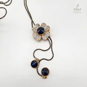 May include: A gold-toned necklace featuring a flower-shaped pendant with clear crystals and a large, dark blue center stone. The necklace has a dark chain and two additional dark blue stones at the bottom. The text "Edited by ESSENCE" is in the upper right corner.