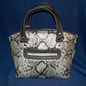May include: A small, silver and black handbag with a snakeskin pattern. The bag has two black handles and a zipper closure.