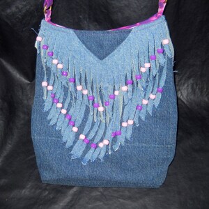 May include: A denim handbag with a fringe design and purple and pink beads. The bag has a v-shaped opening and a purple and pink fabric lining.
