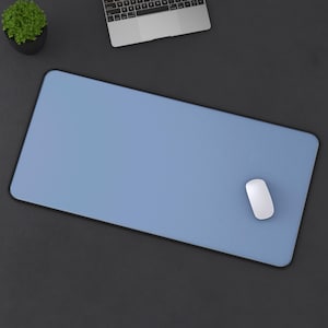 May include: A light blue rectangular mouse pad with a black border. A white computer mouse is on the right side of the pad. A laptop and a small potted plant are in the background.