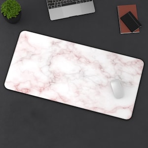 May include: A rectangular mouse pad with a white and pink marble pattern. The mouse pad has a black border and a white computer mouse is on the right side. A laptop, a small plant, and a notebook are also visible.