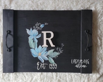 Könnte beinhalten: Schwarzes Holztablett mit einem weißen, bemalten Blumendesign, dem Buchstaben "R" und dem Text "Est. 1999" und "Ladybug Artisan".