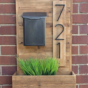 Puede incluir: Un conjunto de buzón y jardinera de madera para pared, con un buzón de metal marrón oscuro, una placa vertical con los números negros 721 y una jardinera rectangular de madera que contiene hierba artificial verde.