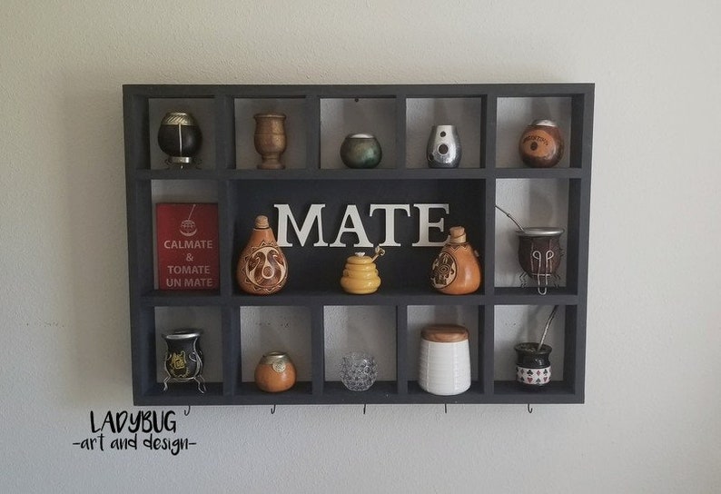 K&ouml;nnte beinhalten: Ein schwarzes Holzregal mit mehreren F&auml;chern, das verschiedene Mate-K&uuml;rbisse, Tassen und Zubeh&ouml;r zeigt. Das Wort "MATE" ist in wei&szlig;en Buchstaben auf dem Regal geschrieben. Das Regal ist an einer wei&szlig;en Wand montiert.