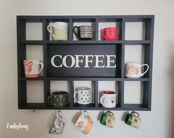 May include: Black wooden coffee mug rack with hooks on the bottom. The rack has 12 compartments and a sign that says "COFFEE". There are various mugs in the compartments and hanging from the hooks.