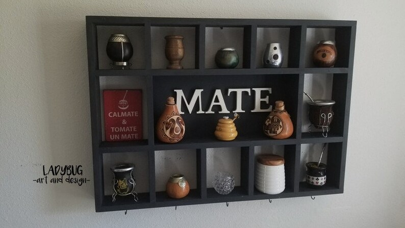 K&ouml;nnte beinhalten: Ein schwarzes Holzregal mit 16 quadratischen F&auml;chern, in denen verschiedene Mate-K&uuml;rbisse, Tassen und Zubeh&ouml;r ausgestellt sind. Das Wort "MATE" ist in wei&szlig;en Buchstaben auf dem Regal geschrieben. Das Regal ist an einer wei&szlig;en Wand montiert.