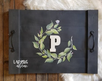 Könnte beinhalten: Ein schwarzes Holztablett mit schwarzen Metallgriffen. Das Tablett ist mit einem grünen Blätterkranz und einem weißen Buchstaben "P" in der Mitte bemalt. Der Text "Ladybug Artisan" ist in weiß in der linken unteren Ecke des Tabletts geschrieben.