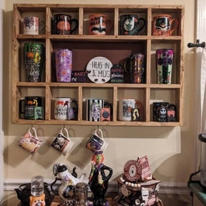 K&ouml;nnte beinhalten: Ein Holzregal mit mehreren F&auml;chern zeigt eine Sammlung von Halloween-Tassen. Die Tassen haben verschiedene Designs, darunter K&uuml;rbisse, Flederm&auml;use, Hexen und Geister. Das Regal ist mit einem Schild verziert, auf dem "Hug in a Mug" steht.