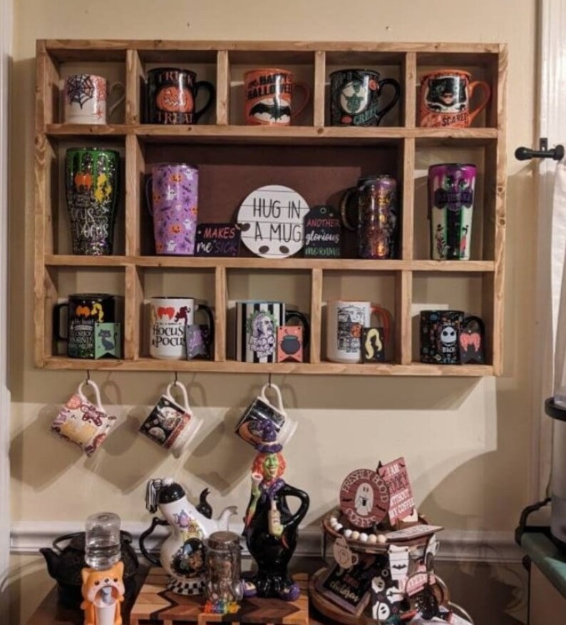 K&ouml;nnte beinhalten: Ein Holzregal mit mehreren F&auml;chern zeigt eine Sammlung von Halloween-Tassen. Die Tassen haben verschiedene Designs, darunter K&uuml;rbisse, Flederm&auml;use, Hexen und Geister. Das Regal ist mit einem Schild verziert, auf dem "Hug in a Mug" steht.