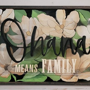 Puede incluir: Un letrero de madera con un marco negro y un inserto de acrílico transparente. El inserto de acrílico presenta un diseño floral con flores de magnolia blancas y beige y hojas verdes. El texto "Ohana Means Family" está escrito en negro en el inserto de acrílico.