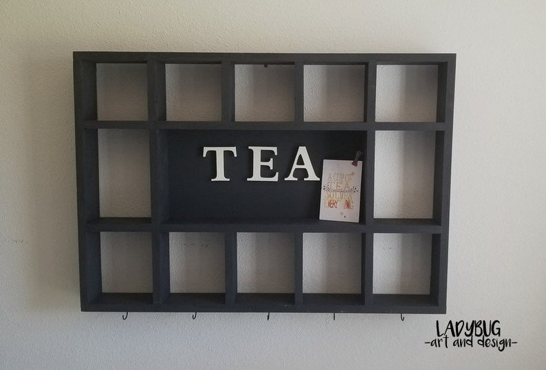 K&ouml;nnte beinhalten: Ein schwarzes Holzregal mit 15 quadratischen F&auml;chern, beschriftet mit "TEA" in wei&szlig;en Buchstaben. Das Regal hat Haken an der Unterseite und ist an einer wei&szlig;en Wand montiert.