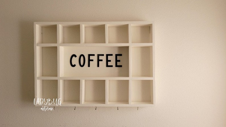 Puede incluir: Estanter&iacute;a de madera blanca con 12 compartimentos y ganchos debajo. La estanter&iacute;a tiene la palabra "COFFEE" en letras negras en la parte delantera.
