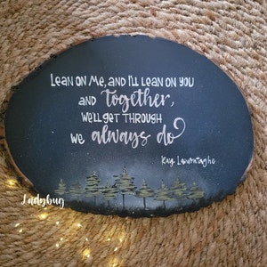 May include: A dark blue wooden sign with a white painted quote: "Lean on me, and I'll lean on you and together, we'll get through we always do." The quote is attributed to Kay Launontaghe. The sign is decorated with a silhouette of a forest.