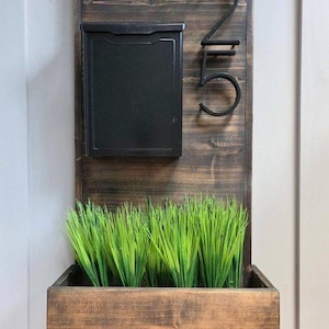 Puede incluir: Un letrero de dirección de madera con un buzón negro, el número 25 y una jardinera llena de hierba verde. El letrero está hecho de madera teñida oscura y está montado en una pared blanca.