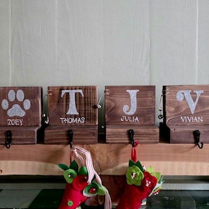 May include: Four rectangular dark brown wooden stocking holders with black metal hooks. Each block features a silver glitter design: a paw print labeled Zoey, and the letters T, J, and V above the names Thomas, Julia, and Vivian respectively.