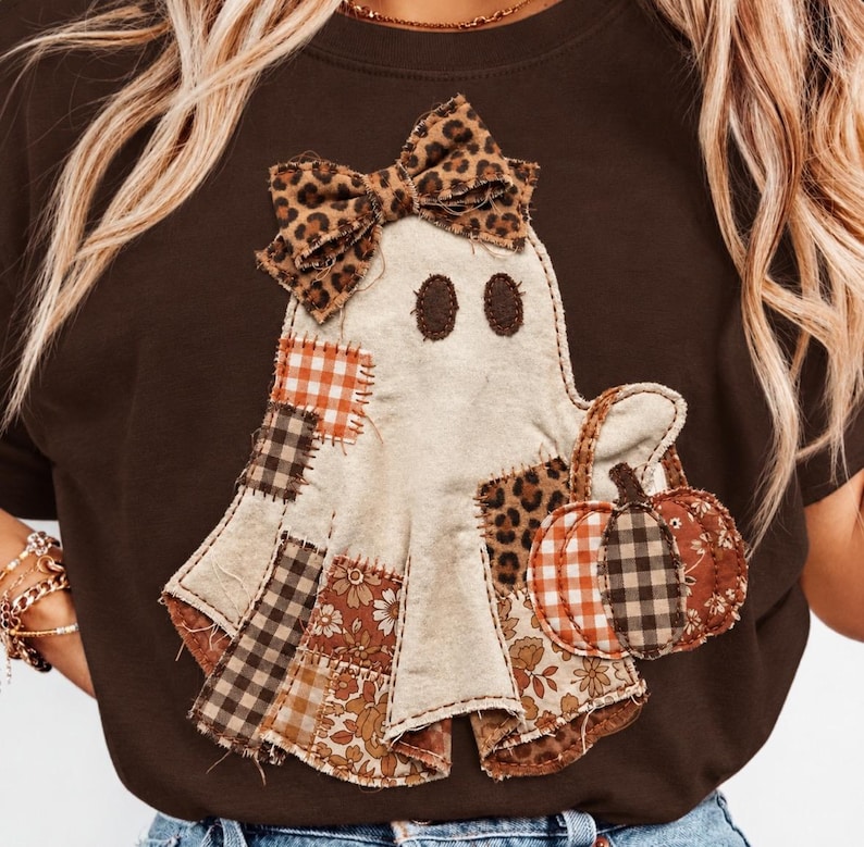 May include: A dark brown t-shirt featuring a central ghost appliqu&eacute; made from various fabric patches. The ghost has a leopard print bow, dark oval eyes, and holds a pumpkin composed of orange gingham, floral, and brown plaid fabric segments.