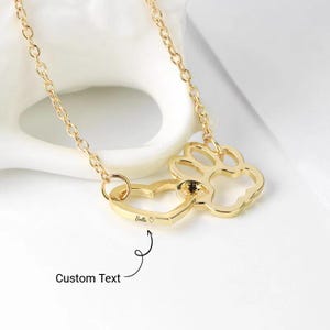 May include: A gold-colored necklace featuring a heart and paw print pendant. The heart-shaped pendant has the name "Bella" engraved on it. The necklace is on a white surface, with the words "Custom Text" in the image.