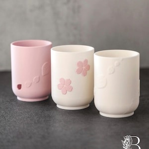 Peut inclure: Trois tasses en céramique, de forme cylindrique. L'une est rose clair, les autres sont blanc cassé. La tasse rose a des motifs en forme de feuilles. Les tasses blanc cassé ont des motifs floraux roses. Elles sont posées sur une surface sombre.
