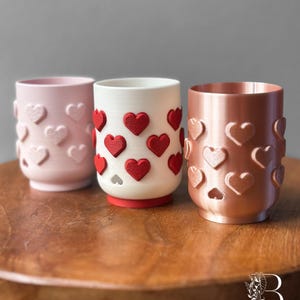 May include: Three cylindrical candle holders in pink, white, and copper colors, each adorned with heart-shaped cutouts and raised heart accents. The candle holders are arranged on a wooden surface, showcasing a Valentine's Day theme.