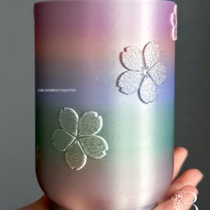 May include: A cylindrical tumbler with a gradient finish of pink, purple, blue, and green. The tumbler features embossed silver cherry blossom designs. The text "@BLOOMBOUTIQUEPDX" is visible.