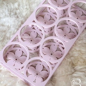 May include: A light pink, rectangular tray with a floral design. The tray features circular cutouts, each containing a stylized flower. The flowers have five petals and a star-shaped center. The tray is made of a solid material and sits on a fluffy, white surface.