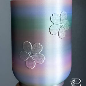 May include: A cylindrical planter featuring a smooth gradient finish that transitions through shades of pink, purple, blue, and green. Two embossed cherry blossom flower motifs are positioned on the exterior surface. The text BLOOMBOUTIQUEPDX is visible.