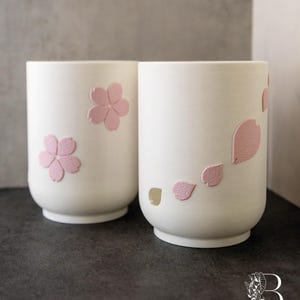 May include: Two white ceramic cups with pink cherry blossom designs. One cup features a full flower, while the other has petals. The cups are cylindrical with a slightly rounded base, perfect for tea or coffee.