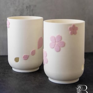 以下が含まれることがあります： ピンクの桜の花模様が施された、白いセラミックカップ2つ。1つのカップにはピンクの花びらがいくつかあり、小さな穴があります。もう1つのカップにはピンクの桜が3つあります。カップは円筒形で、暗い表面に置かれています。