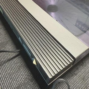 May include: A silver and black vintage turntable with a clear, purple-tinted platter. The turntable has a rectangular design with a series of parallel lines on the front. A black power cord is visible.