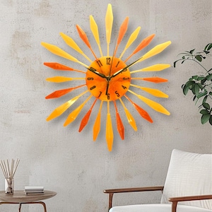 Puede incluir: Reloj de pared decorativo con diseño de rayos de sol. La esfera es naranja con números y manecillas negras. Los rayos son una gradación de amarillo y naranja. El reloj está montado en una pared gris.